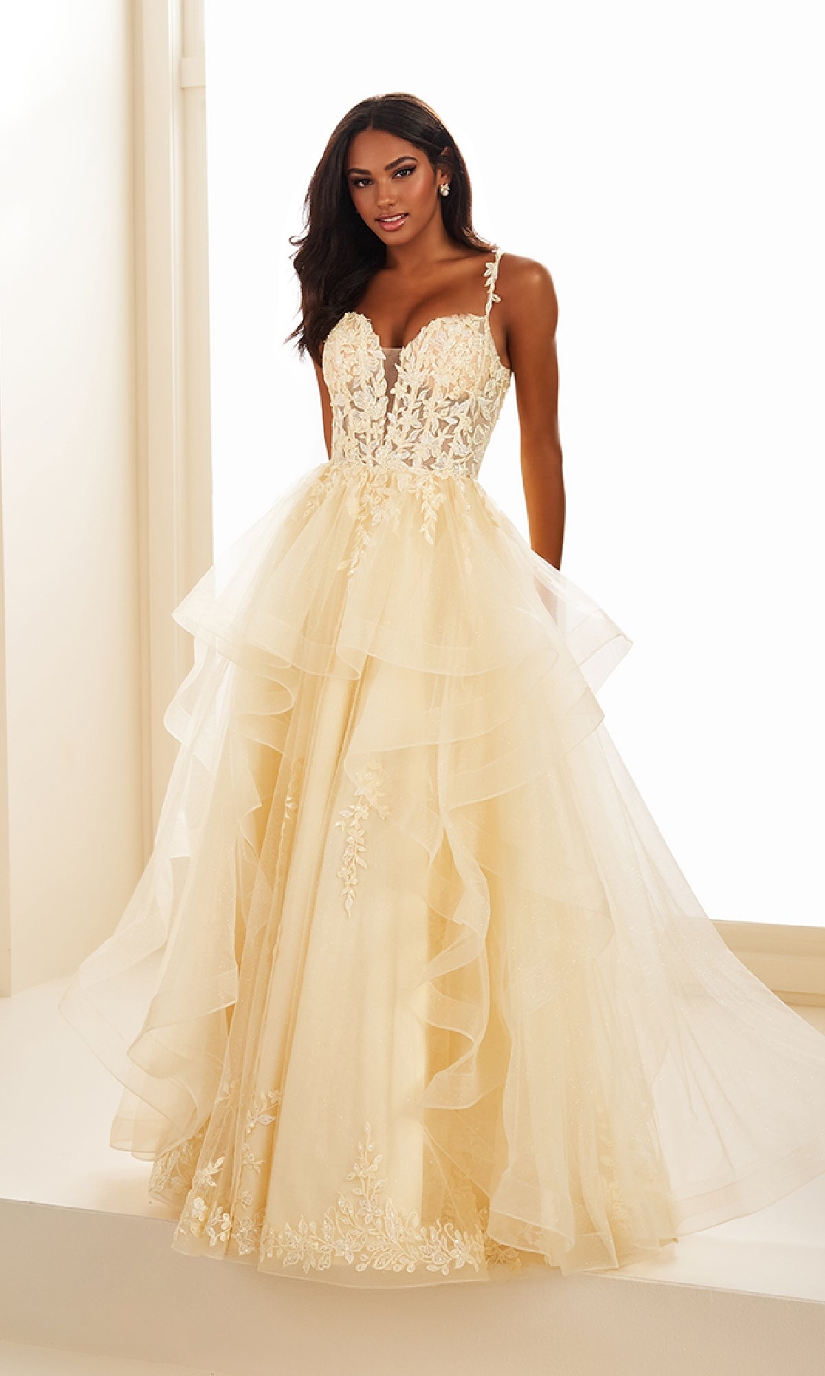 A woman poses indoors against a light background wearing the Ellie Wilde EW36081 long prom dress, featuring a cream-colored, layered glitter tulle gown with lace detailing and a fitted bodice.